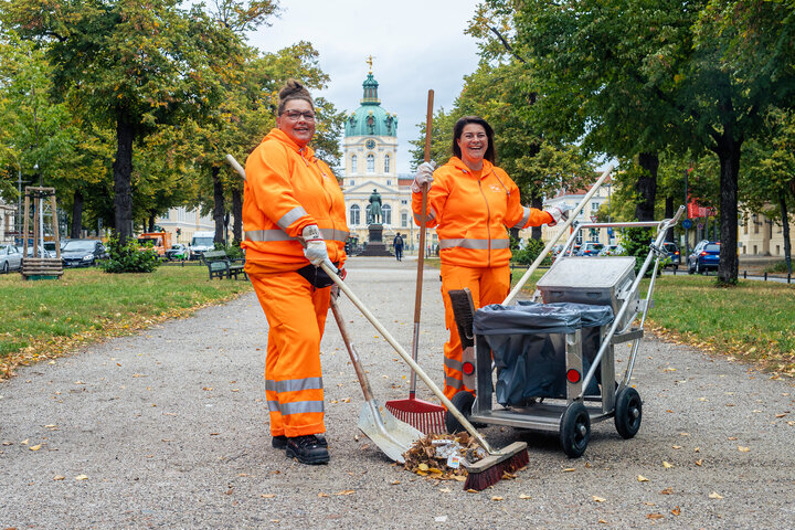 zwei Frauen in orangefarbener Kleidung aus der Straßenreinigung der BSR mit Besen und Kehrichtkarre